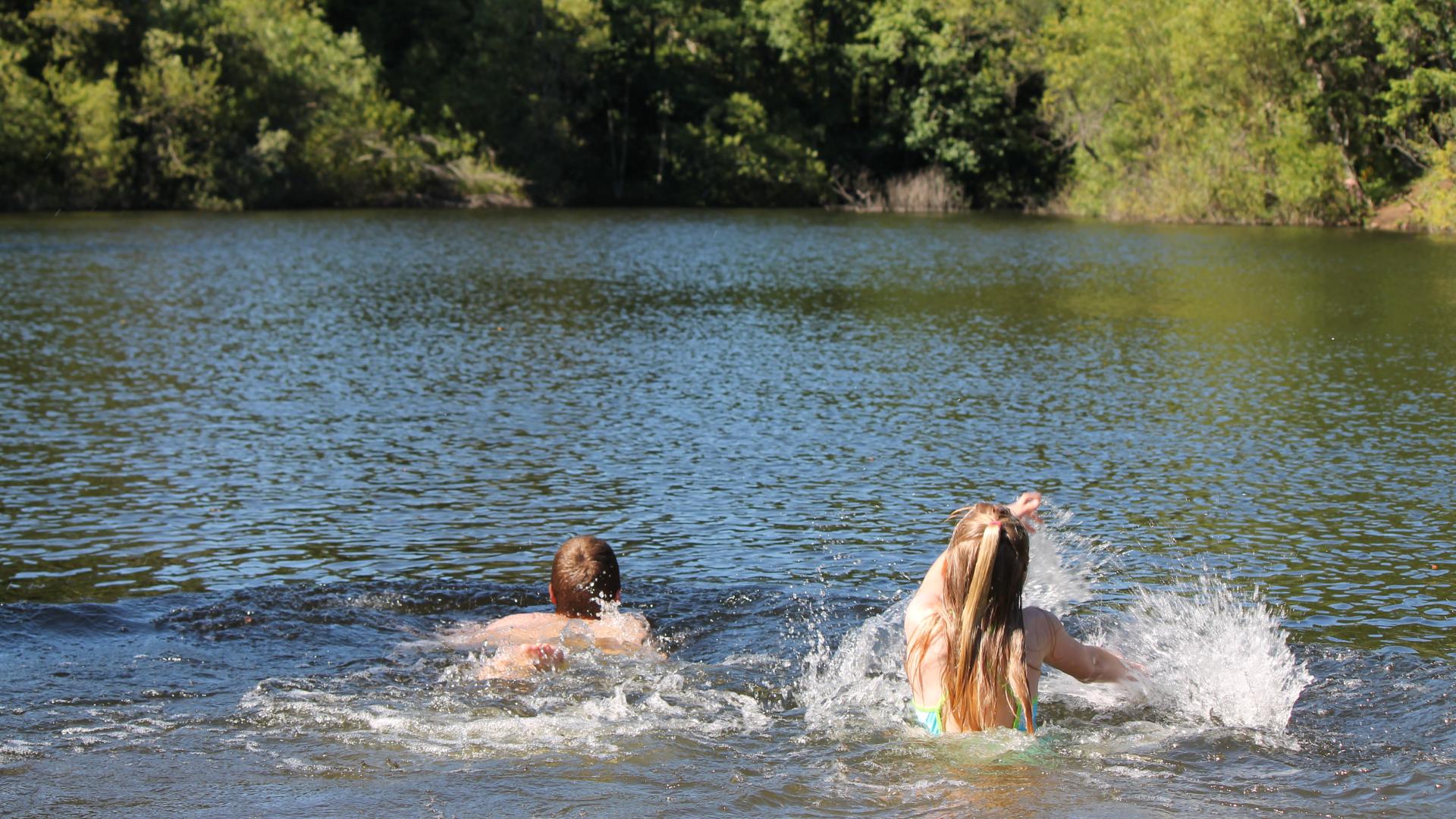 Two children swimming and splashing in a lake surrounded by green trees.