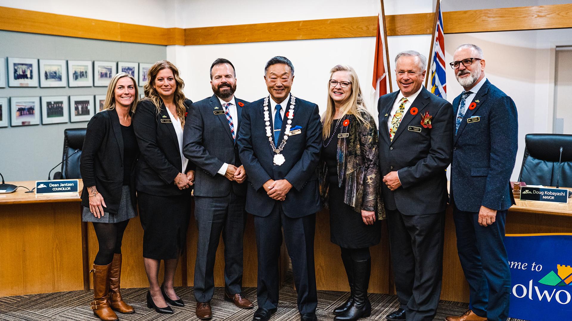 Group of city council members, including the mayor, standing together in a council chamber with flags and framed photos in the background.