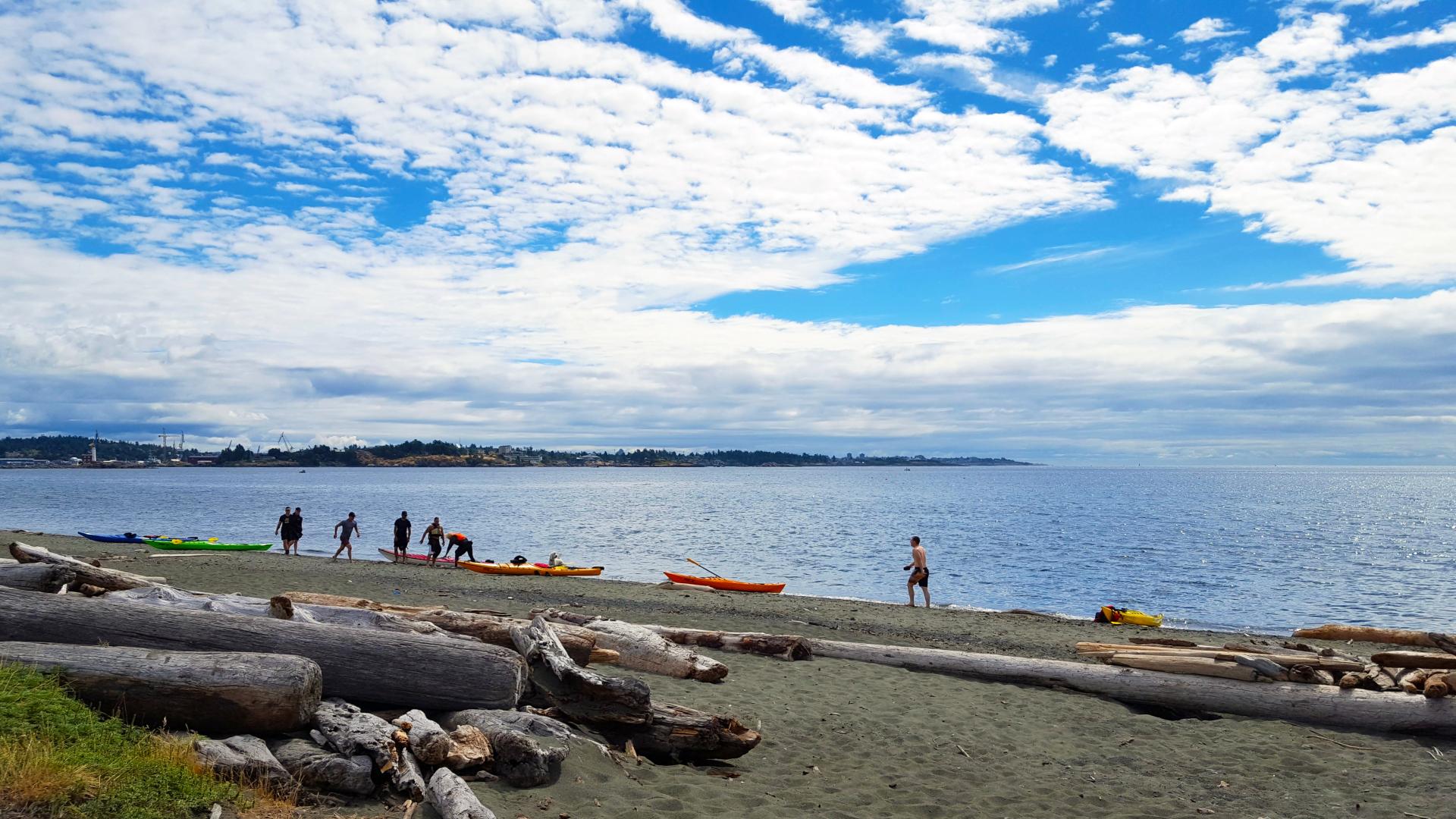 Beach with driftwood and people preparing kayaks on the shore under a partly cloudy sky.