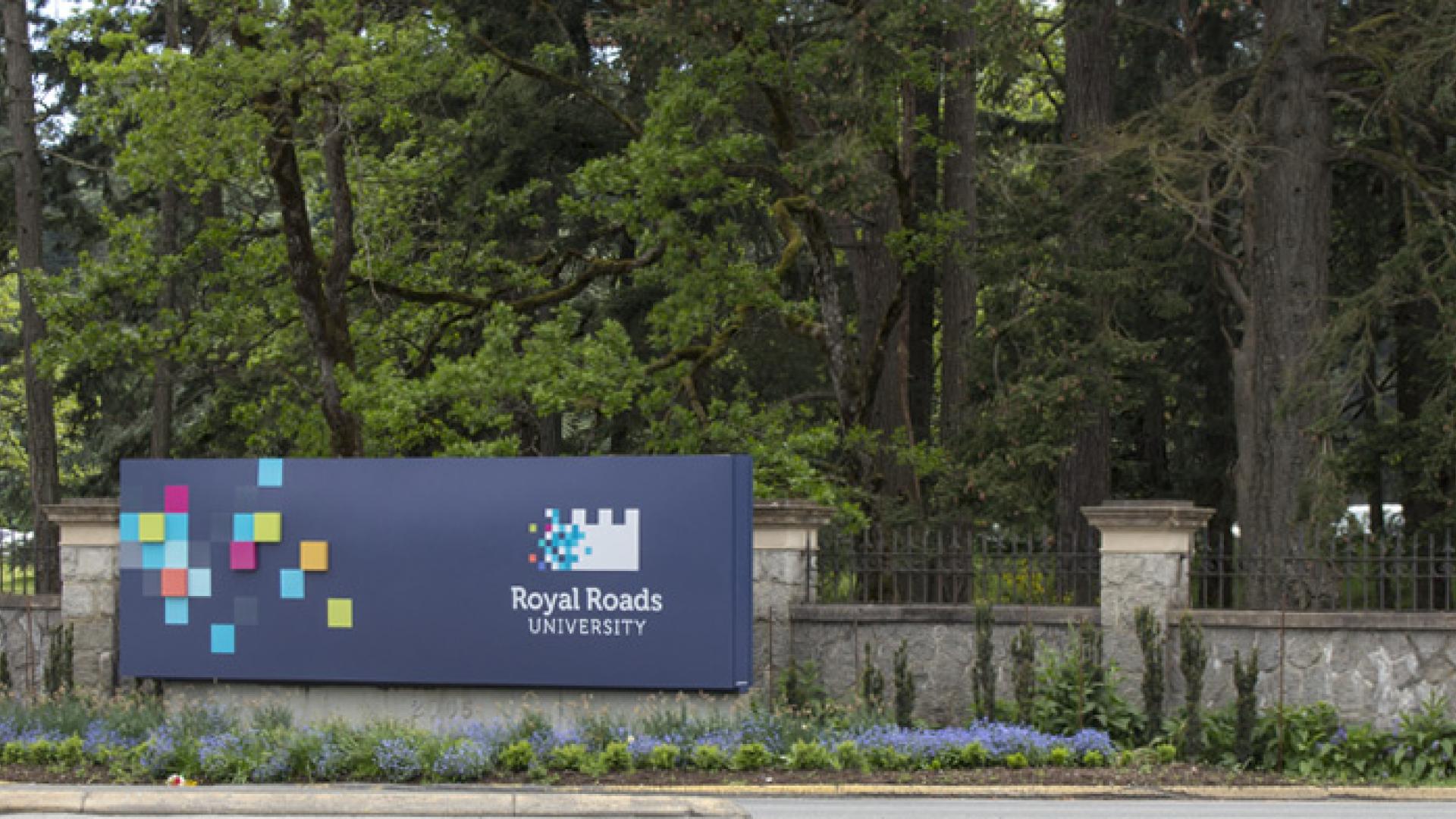 Sign for Royal Roads University in front of a wooded area with colorful square graphics.