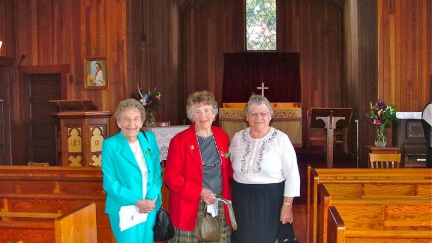 Three elderly women inside of a church. They are flanked by pews and the altar is in the background.