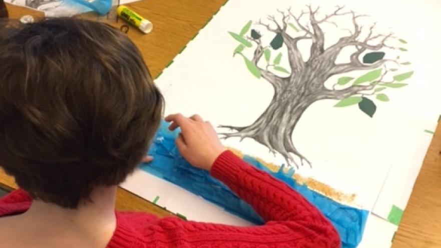 Person with a red sweater working on a multi-media piece of art. The artwork has a the drawing of a tree on it.