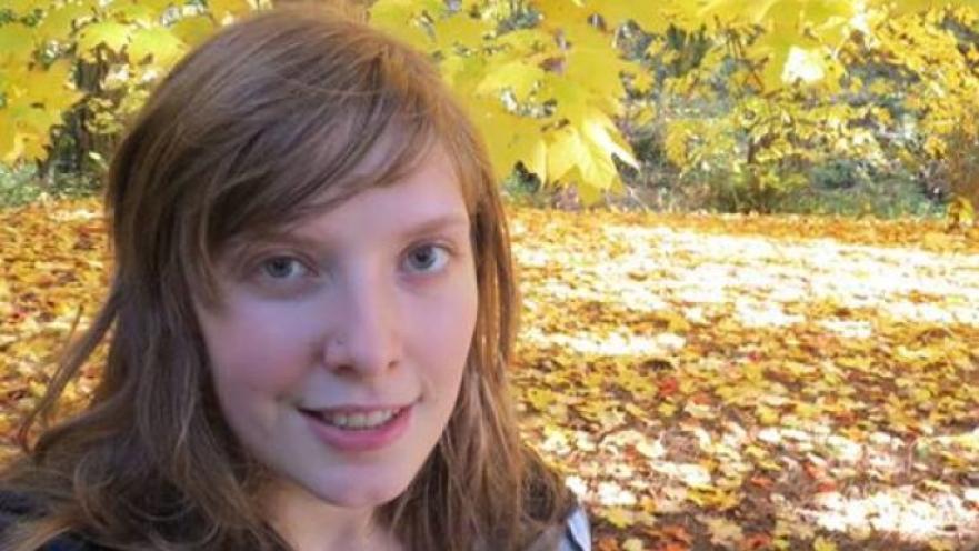 Selfie of woman with long brown hair. There are fallen leaves on the ground and trees with yellow leaves.