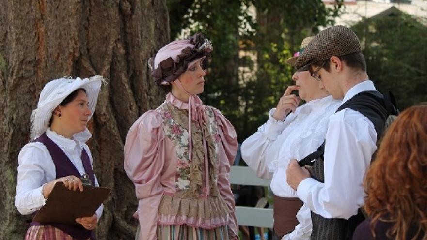 Group of people standing in front of tree. The subjects are all wearing old-timey clothes.