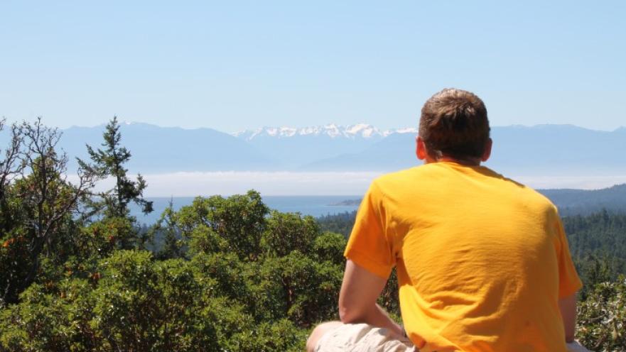 Man wearing bright orange shirt staring off into the distance. There are mountains and a wooded area in the background.