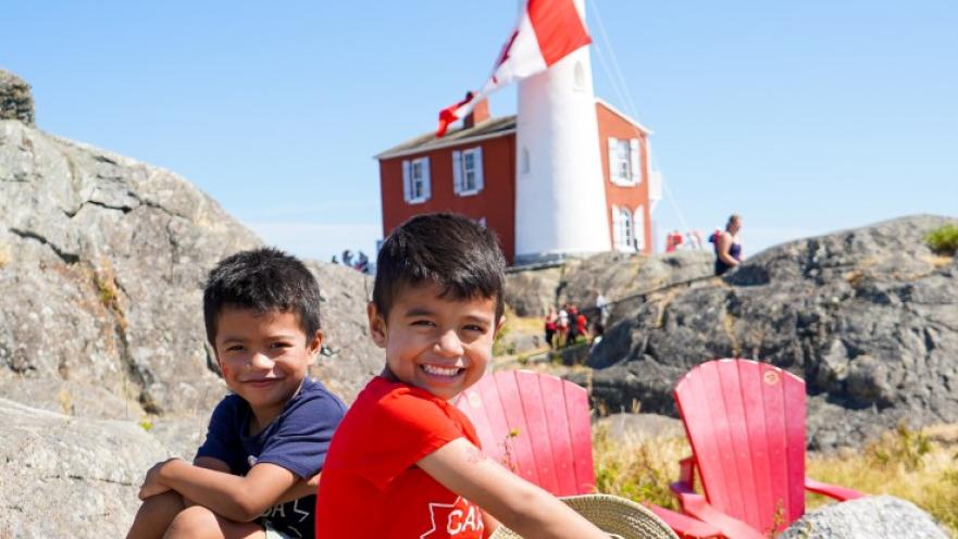 Two children smiling sitting on boulders. There is a lighthouse in the background.