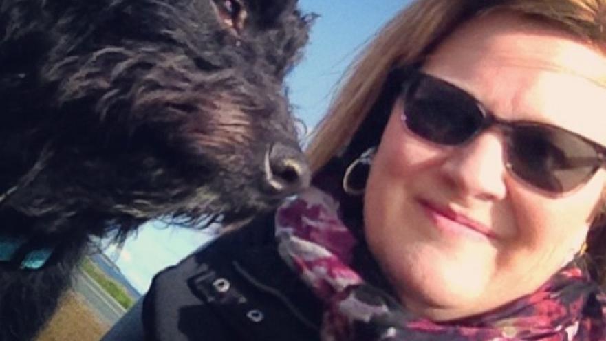 Selfie of woman and their dog. The dog has shaggy, dark fur. The woman is wearing a scarf and dark sunglasses.