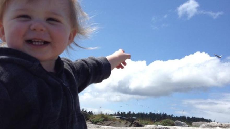 Blonde toddler smiling while pointing at white clouds in the sky. The toddler is at a beach.