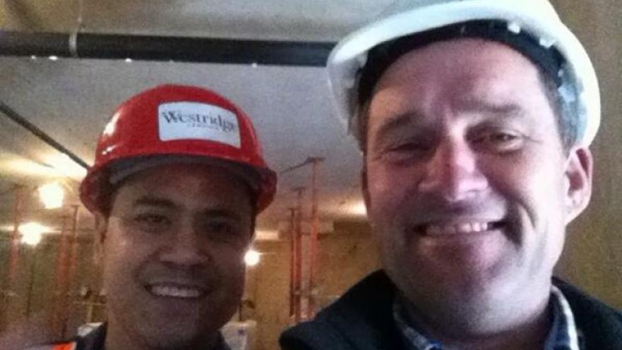 Selfie of construction workers. One worker is wearing a white construction helment while the other is wearing a red construction helmet and a high-vis vest.