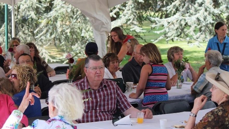 Group of people sitting by large tables. A large, white tent provides shade to the subjects.