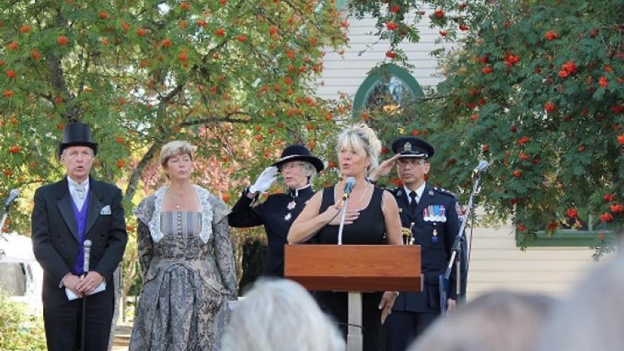 Woman giving a speech at a podium. There are a group of people dressed in old-timey clothes behind the subject.