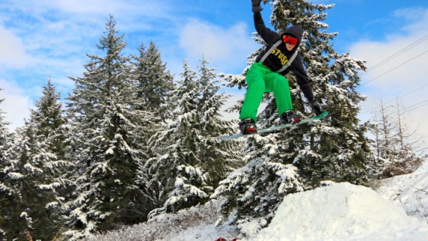 Snowboarder jumping over snow ramp. There are trees covered in snow in the background.