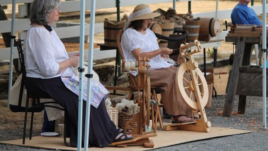 Two women spinning wool into yarn. A white tent offers the subjects some shade.