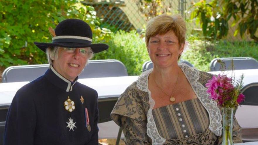 Two women wearing old-timey clothes sitting behind a table.