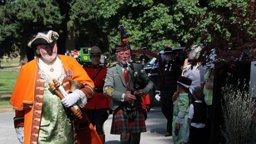 Person dressed as an old-timey town-crier carrying some scrolls. There is a bagpiper following the town crier.