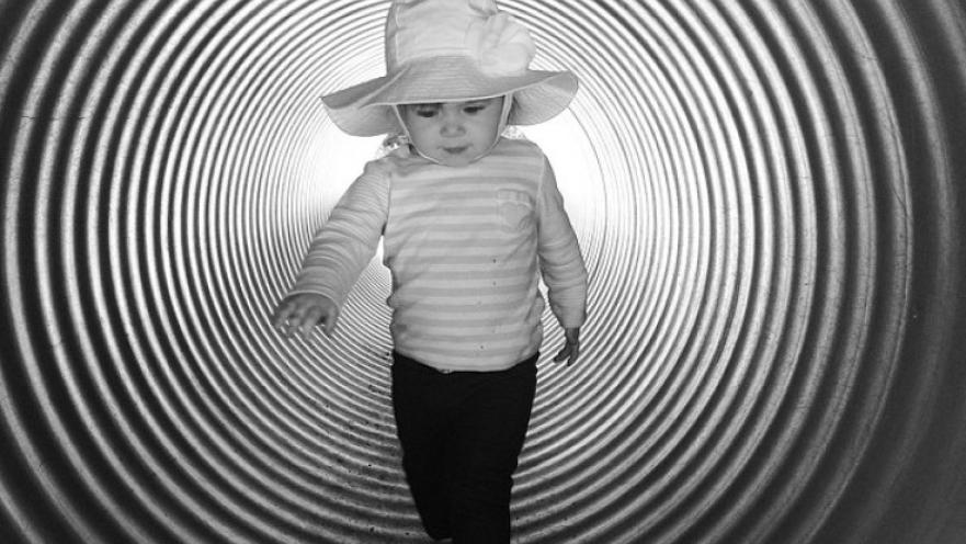 Monochrome photo of child walking down a corrugated pipe. The child is wearing a bucket hat.