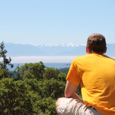 Man wearing bright orange shirt staring off into the distance. There are mountains and a wooded area in the background.