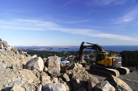 Construction scene showing a Volvo excavator on a rocky hill with a panoramic view of a city and ocean in the background.