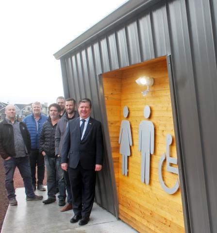 Group of men standing in front of a modern restroom facility with wood paneling and metal siding, featuring signage for men, women, and accessible restrooms.