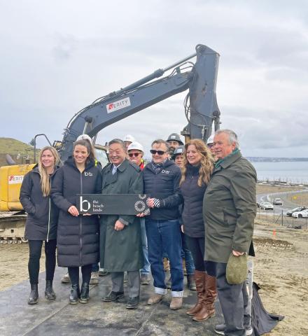 Group photo of people at a construction site holding a sign for The Beach Lands, with an excavator and the ocean in the background.