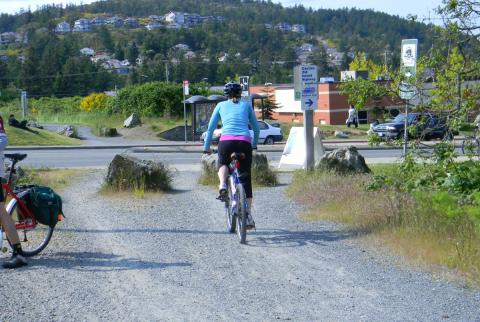 Cyclist in blue jacket and black shorts riding on a gravel path towards a main road, with hills and houses in the background.