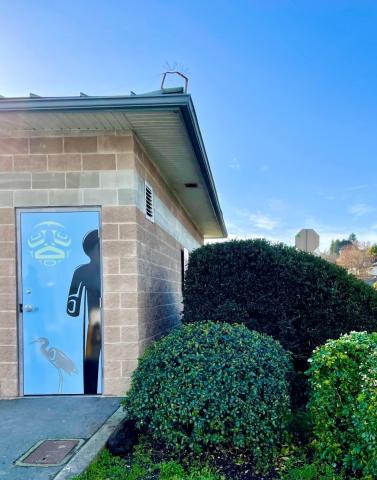 A small building with a door featuring a heron and First Nations art, surrounded by greenery.