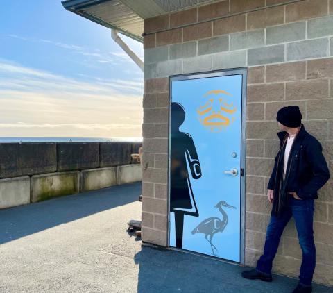 A man standing beside a building with an ocean view, the door decorated with a heron and First Nations art.