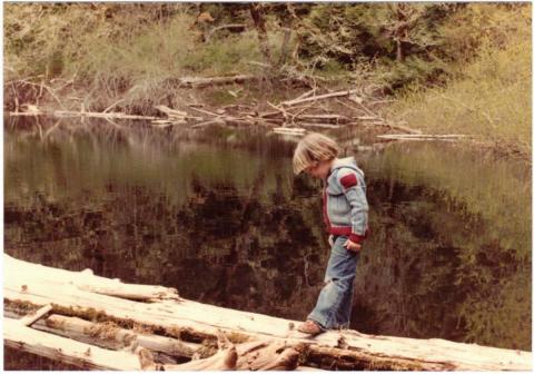 A young child in a denim jacket walking on a log near a calm pond with trees and shrubs in the background.