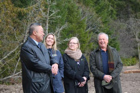 A group of four people standing outdoors, talking and smiling, with trees in the background.