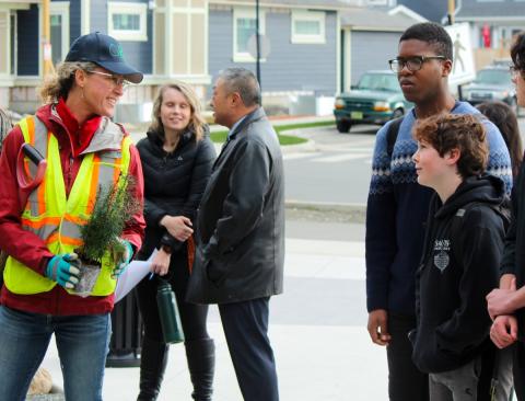 A woman in a reflective vest holding a small tree, talking to a group of people in an urban setting.