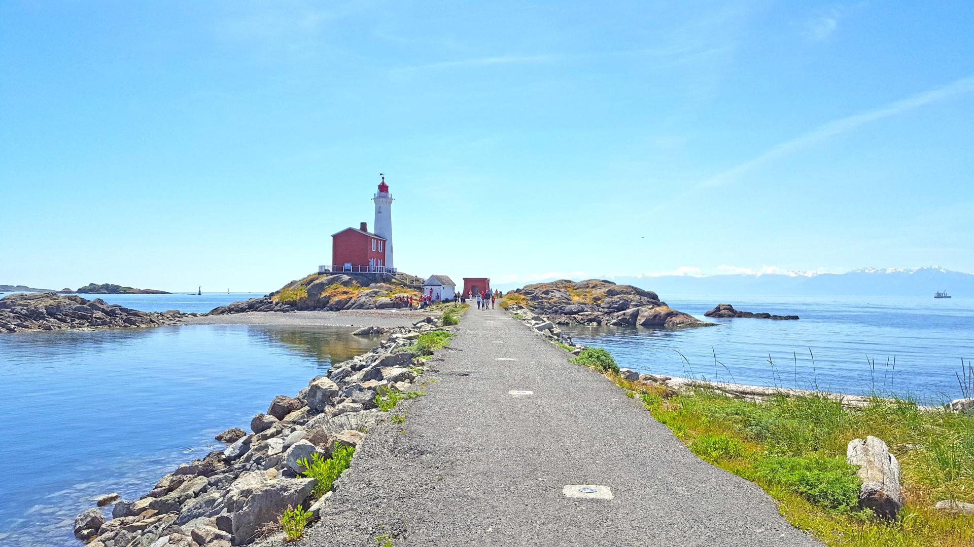 A scenic view of a lighthouse on a small island, connected to the mainland by a narrow rocky causeway, with calm blue waters and a mountainous backdrop.