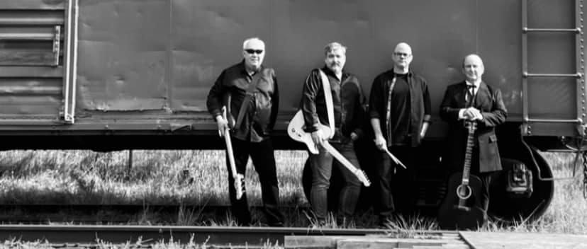 Black and white photo of four men with musical instruments standing in front of a train car. The group includes two guitarists, a bassist, and a drummer.