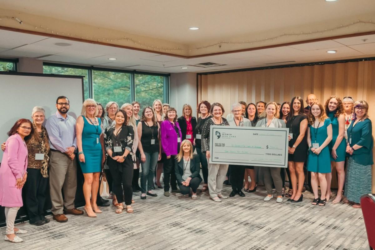 Group of women and a few men standing in a conference room holding a large check for $7,900 from the organization Women Who Care.