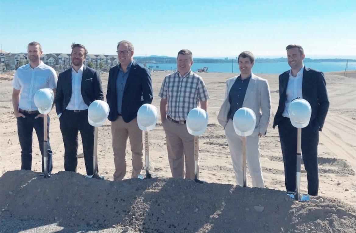 Six men dressed in business casual attire stand behind shovels and hard hats at a groundbreaking ceremony on a sunny day near the ocean.