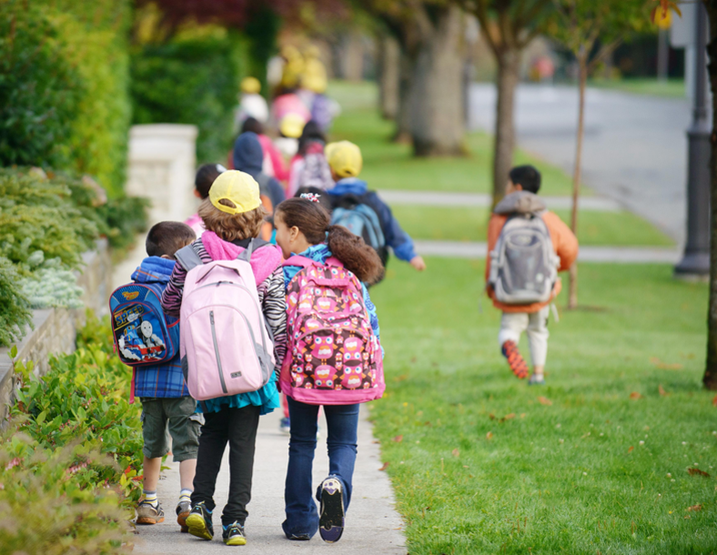 Children walking to school with backpacks on a tree-lined sidewalk.