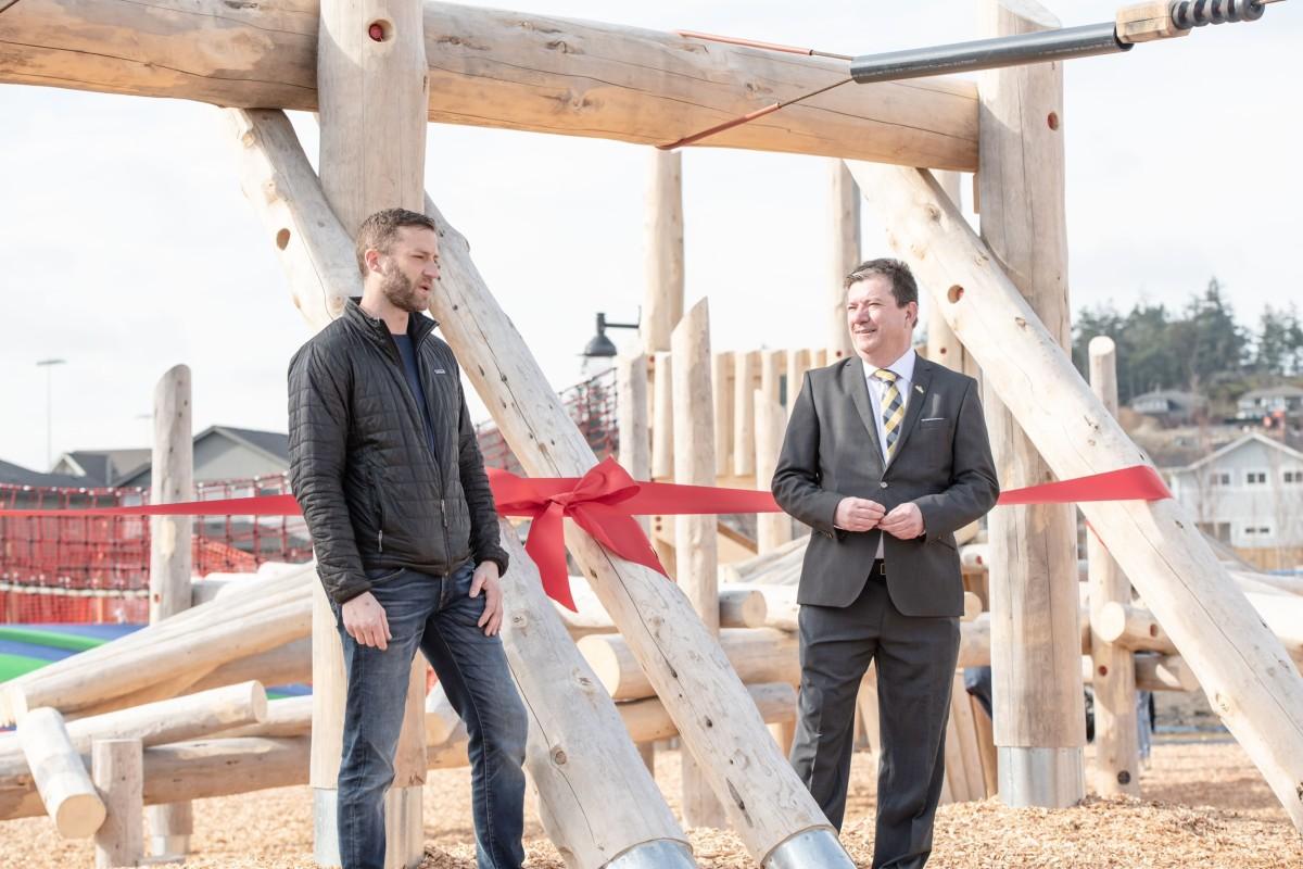 Two men standing in front of a wooden playground structure with a red ribbon for a ribbon-cutting ceremony.