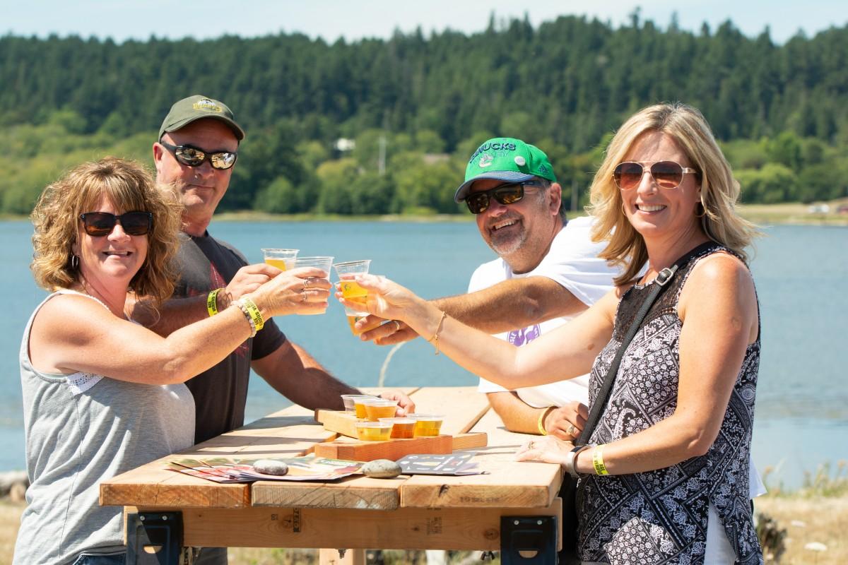 Four people toasting with drinks by a lake, enjoying a sunny day.