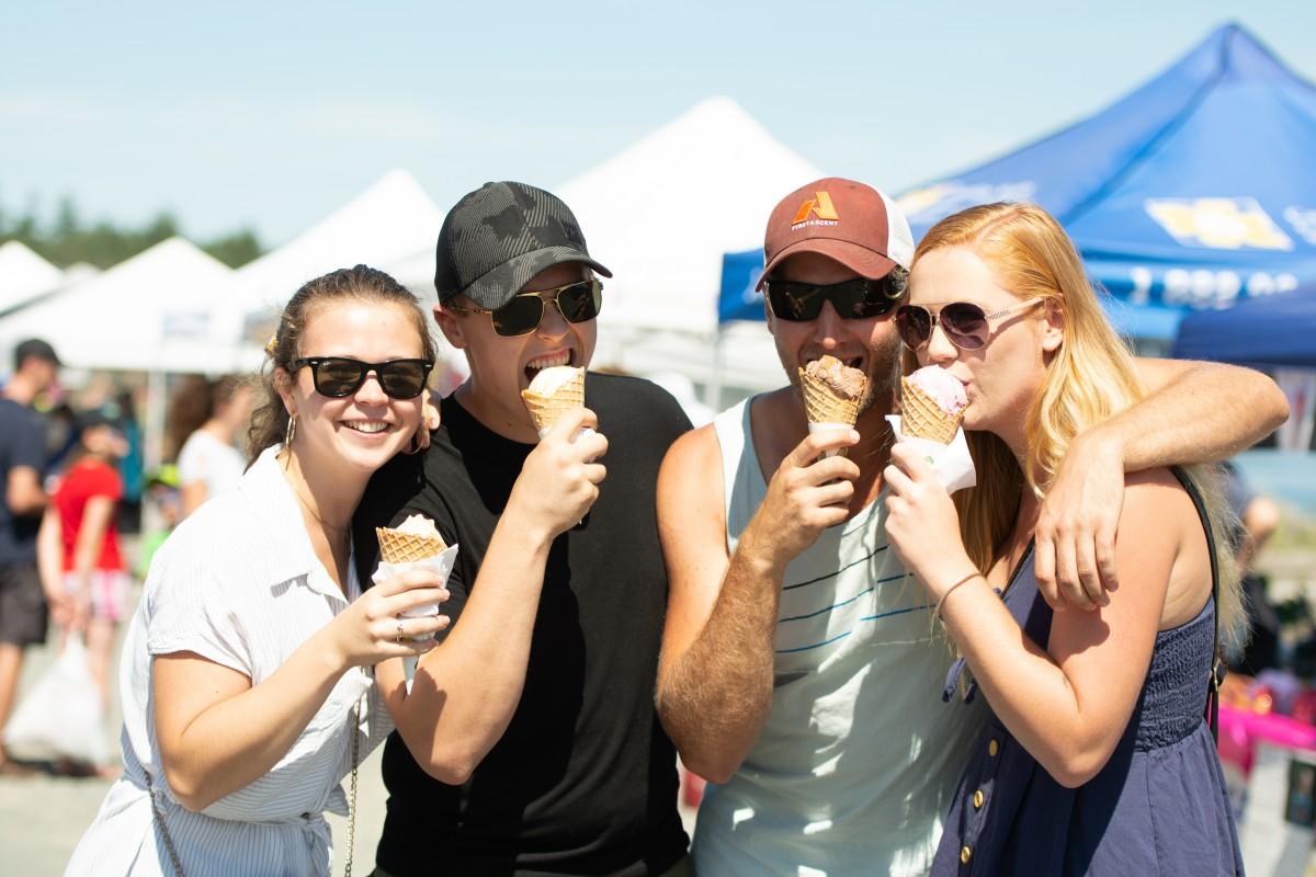 Four friends smiling and eating ice cream cones at an outdoor event.