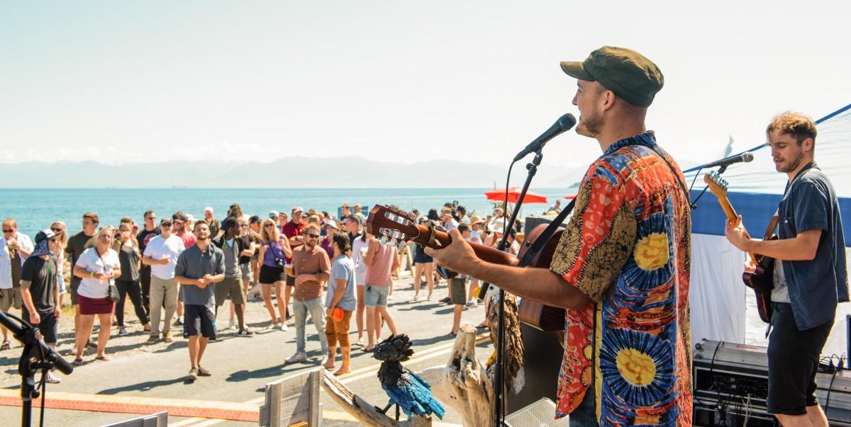 Musicians performing at an outdoor event with a crowd and ocean backdrop.