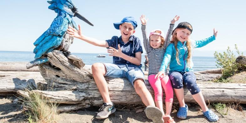 Three children sitting on a log with a blue bird sculpture at the beach, smiling and posing.