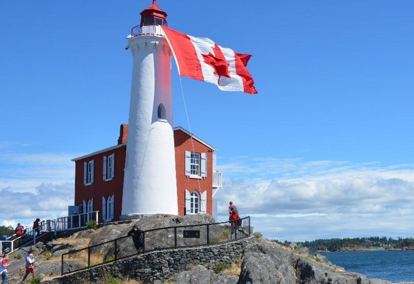 White lighthouse with red top and attached brick building, flying large Canadian flag, on rocky coast with visitors.