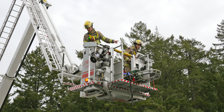 Two firefighters in a ladder truck performing an operation, surrounded by tall trees.