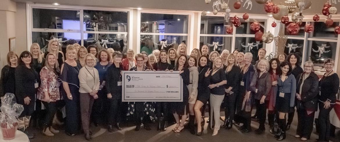 Group photo of women holding a large check at a Women Who Care event, with festive decorations in the background.