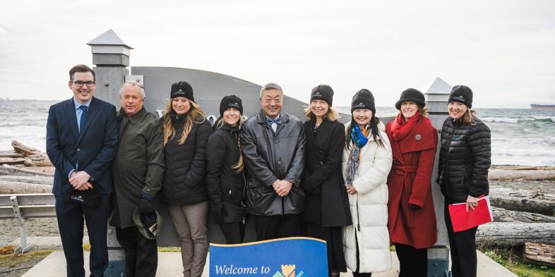 A group of nine people dressed warmly, posing in front of a sign on a windy beach.