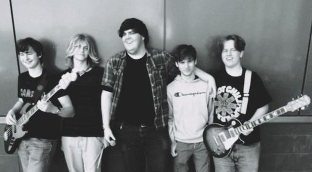 Black and white photo of five young men in a band, standing together with musical instruments. Two have guitars, one has a bass, and one is holding a microphone.