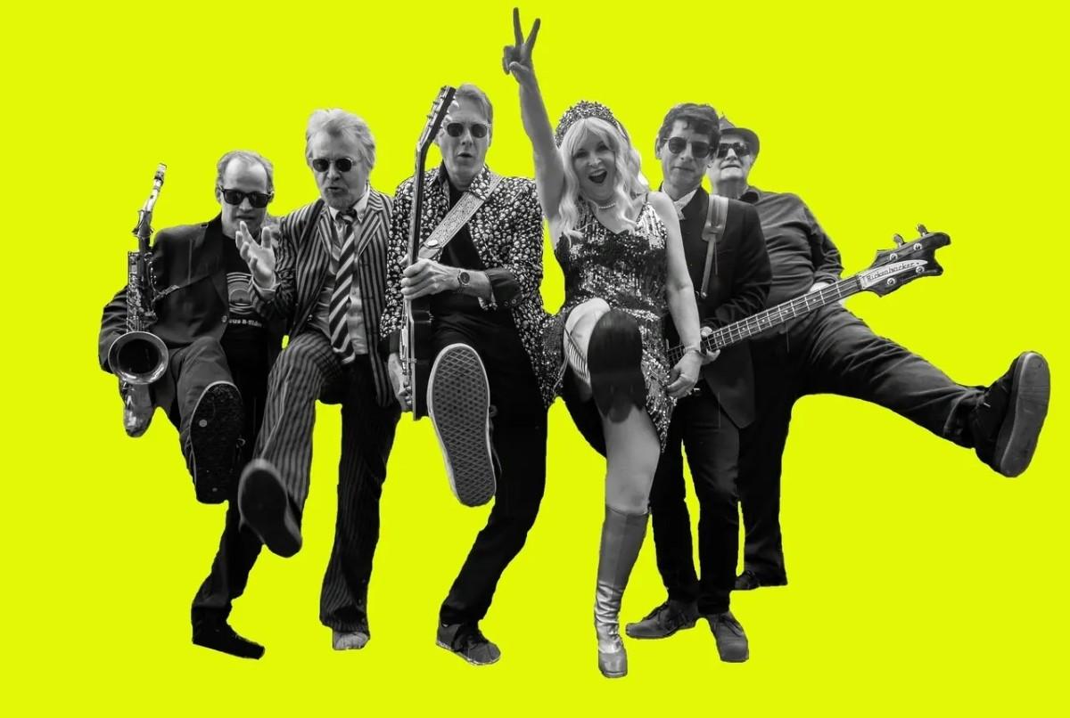 Black and white photo of a lively band against a bright yellow background. The members are mid-kick, with a saxophone, guitars, and a female vocalist in the centre.