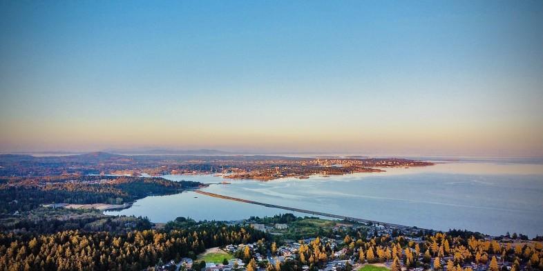 Aerial view of a coastal landscape at sunset, featuring a mix of urban and natural areas. Dense forests and residential neighborhoods are in the foreground, with a body of water and a long, narrow land strip leading to a distant city skyline.