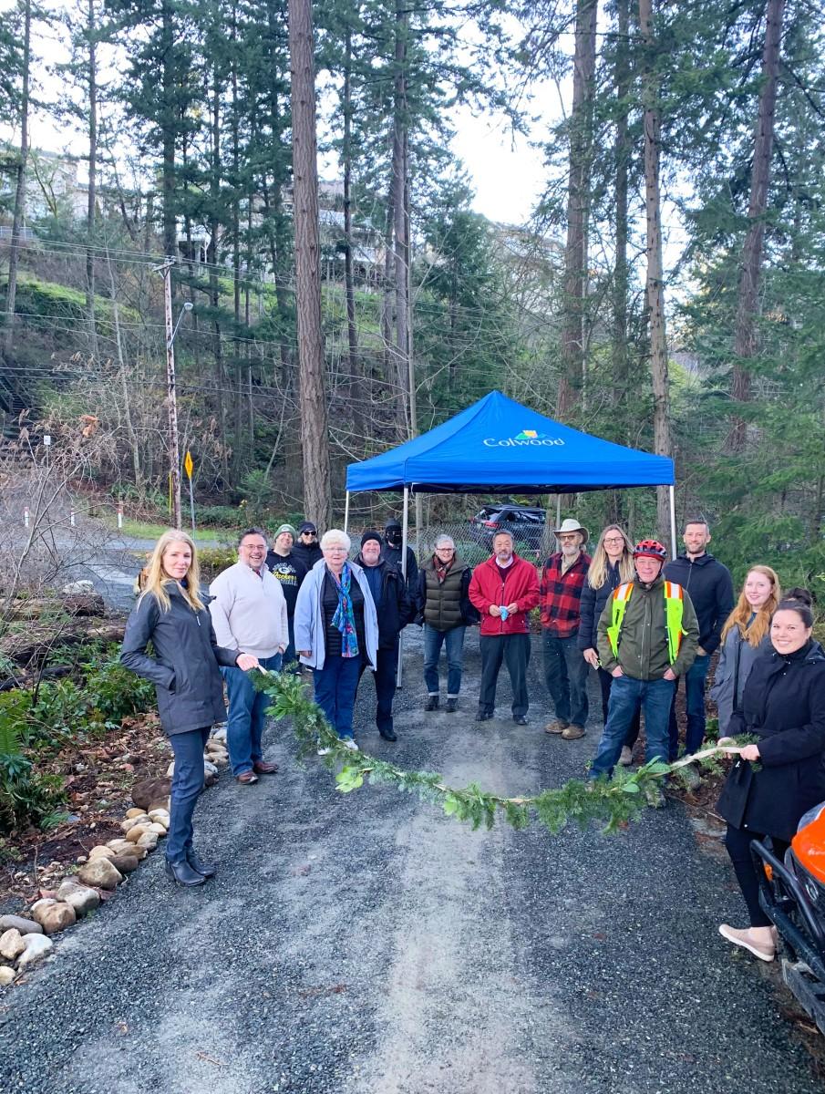 Group of people, including community members, gathered under a Colwood tent for a ribbon-cutting event in a wooded area.