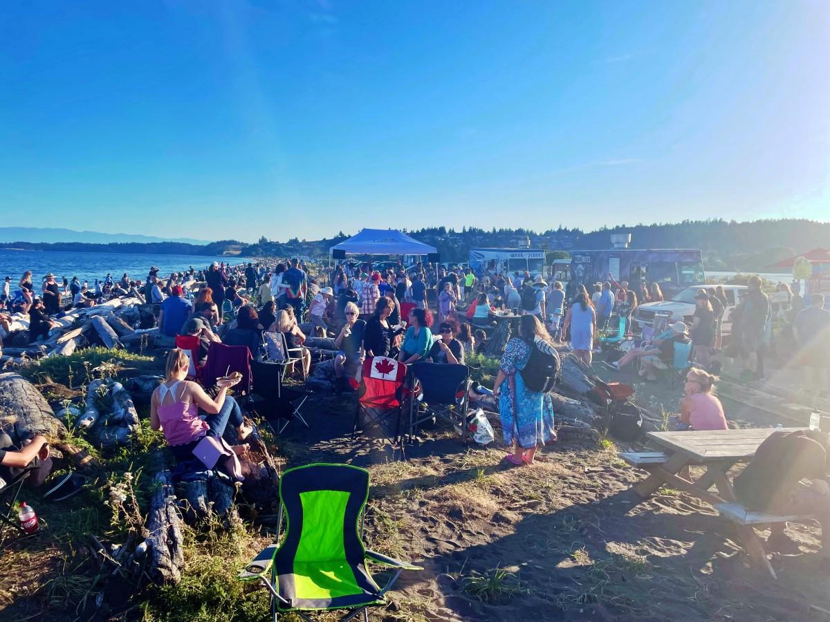 Large crowd of people gathered on the beach, sitting on chairs and logs, with food trucks and a Colwood tent in the background.