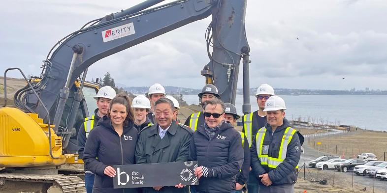 Group of people posing with a sign for The Beach Lands in front of an excavator at a construction site, overlooking the ocean.